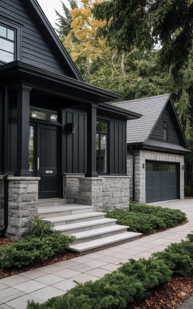 A modern black house features smooth black siding contrasted by rough-cut gray stone accents covering the foundation and vertical pillars along the front. The main entrance is defined by a solid black door with a brushed silver door handle and a rectangular window positioned to the side. A detached stone garage sits to the right of the house, with a single dark gray garage door matching the house’s aesthetic. Low-lying evergreen shrubs and wide stone steps lead to the front door, softening the sharp lines of the architecture and integrating the house into the surrounding landscape.