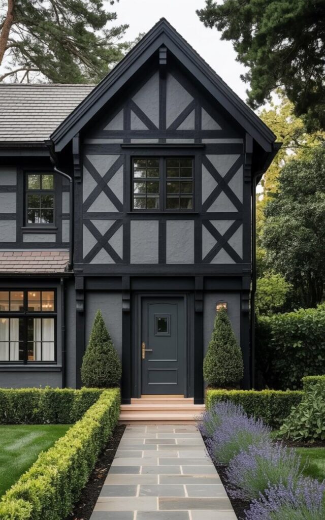 A two-story house with a steeply pitched roof exhibits dark gray timber framing against smooth black stucco walls. Prominent black-painted beams are arranged in a diamond pattern across the façade, while rectangular windows are trimmed with narrow black frames. The front door, featuring a bronze door handle and a small rectangular window, is painted dark gray and recessed slightly within the timber framing. A flagstone pathway leads to the front door, bordered by neatly trimmed boxwood hedges and lavender plants along the foundation.