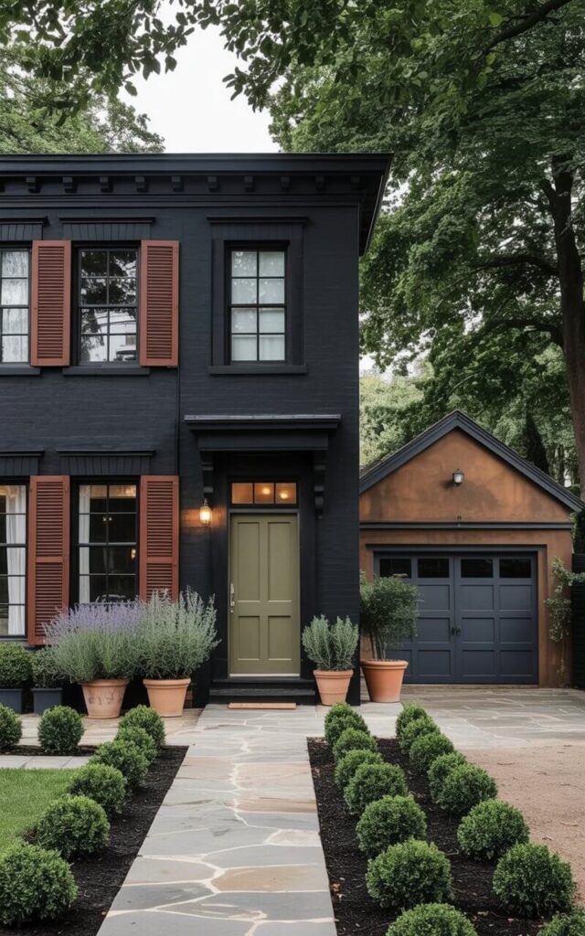 A black house exterior accented with earthy tones, forming a very well designed home with a minimalist yet warm approach. Terracotta pots with plants, rust-colored shutters, and olive-green door panels provide visual interest against the bold black siding. The front door has a door handle and is integrated seamlessly into the minimalist façade. Windows feature thin black frames, while subtle lighting highlights architectural lines. The landscaping is understated, with natural stone walkways and low shrubs, reinforcing the minimalist aesthetic. This black house exterior combines drama, warmth, and carefully curated details for a refined, modern appeal. An earthy garage on the right is clear